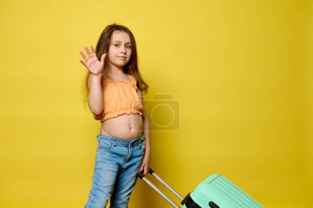 Adorable little traveler girl carrying blue suitcase, waving hello with her hand, looking at camera, isolated over yellow studio background. Copy advertising space. Childhood. Travel. Summer holidays
