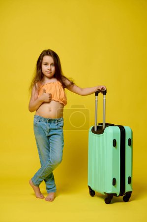 Téléchargez les photos : Portrait vertical pleine longueur d'une petite fille authentique caucasienne, petit enfant de 6 ans en tenue estivale avec valise, montrant un pouce levé, regardant la caméra, isolé sur fond de studio jaune - en image libre de droit