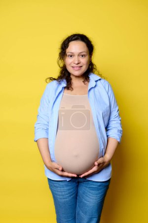 Authentic emotional portrait of a mid adult pregnant woman holding her big belly, smiling looking at camera, isolated yellow background. Pregnancy. Maternity leave. Women's fertility and health