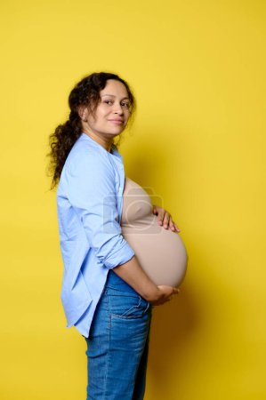 Side portrait of a happy pregnant woman putting hands on her big belly in last trimester of pregnancy, smiling looking at camera, isolated over yellow studio background. Maternity leave concept