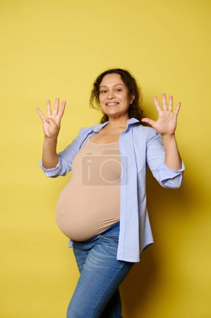 Smiling pregnant woman expecting a baby, showing nine fingers at the camera, isolated on yellow studio background. She's in last trimester of her happy pregnancy. Childbearing. Childbirth. Maternity.