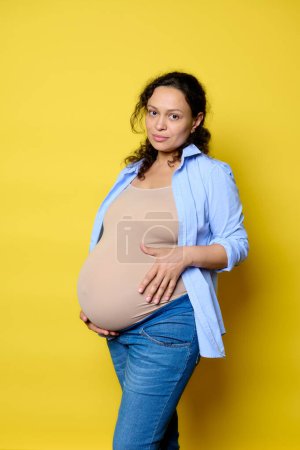 Confident studio portrait of a young pregnant woman in casual denim, touching her belly in last trimester of happy pregnancy, expressing positive emotions looking at camera, isolated yellow background
