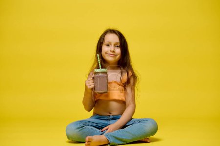 Téléchargez les photos : Petite fille souriante en vêtements d'été, assise sur une pose de lotus, relaxante, rafraîchissante, buvant un cocktail dans une bouteille en verre avec une paille, isolée sur fond de studio jaune. Copier l'espace publicitaire - en image libre de droit