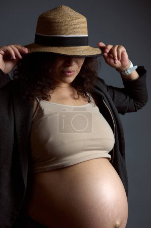 Authentisches Studioporträt einer charmanten sexy schwangeren Frau, werdenden Mutter, mit nacktem Bauch, im letzten Trimester der glücklichen unbeschwerten Schwangerschaft, isoliert über Mode grauen Studiohintergrund.