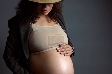 Close-up portrait of young beautiful serene pregnant woman gently caressing her belly, enjoying her happy carefree pregnancy, isolated over fashion gray studio background with free advertising space