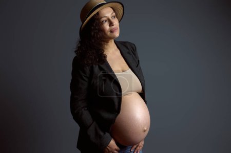 Authentic studio portrait of a multi ethnic attractive and pensive young pregnant woman, expecting baby, holding her beautiful big belly, dreamily looking aside a copy space on fashion gray backdrop