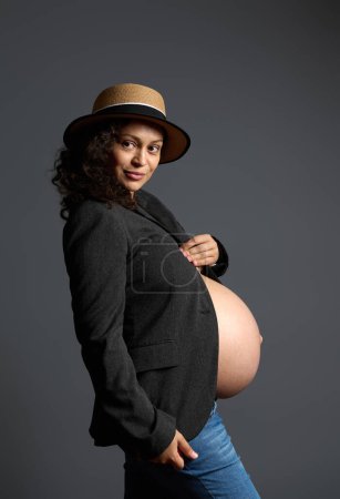 Attractive and pensive young pregnant woman, expecting baby, holding her beautiful big belly, dreamily looking aside a copy ad space on fashion gray backdrop, dressed in gray blazer and blue jeans