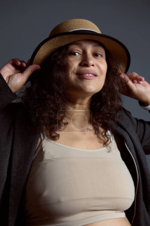 Auténtico retrato de estudio de una mujer embarazada hermosa multiétnica en la parte superior de color beige, chaqueta gris y un sombrero de paja, sonriendo mirando a la cámara, aislado sobre fondo de estudio gris moda