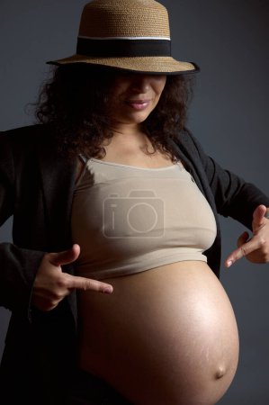 Authentisches Porträt einer sexy Schwangeren, werdenden Mutter, die mit den Fingern auf ihren schönen großen nackten Bauch zeigt, im letzten Trimester der glücklichen Schwangerschaft, isolierter Mode grauer Studiohintergrund.