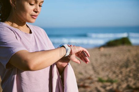 Attractive young sporty female, sportswoman, athlete checking pulse or heart rate mobile app on smart wristwatch, standing on the beach, resting after outdoor workout or morning jog. People and sport