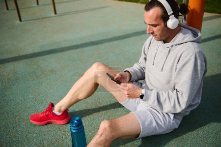 Young active sportsman wearing wireless headphones, checking sports app on his smart mobile phone, sitting in urban sportsground. People and sport. Active healthy lifestyle. Modern technology