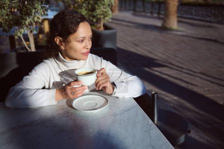 Multi ethnic young stylish woman drinking coffee outdoors at the cafe, dreamily looking aside. Start your day with a boosting cup of energizing coffee drink. People. Lifestyle. Weekend activity