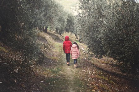 Téléchargez les photos : Vue arrière de deux enfants en vêtements d'hiver chauds marchant le long du sentier forestier bordé d'oliviers dans la vallée de la montagne, entouré par la tranquillité de la nature. Photographie tonique - en image libre de droit