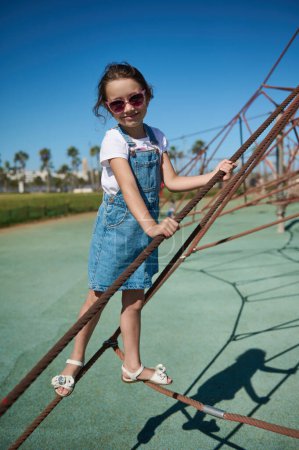 Photo for Little child girl climbing on the climbing net in the urban playground. Full length portrait of cute kid in casual denim playing on the city playground climbing and jumping on the trampoline - Royalty Free Image
