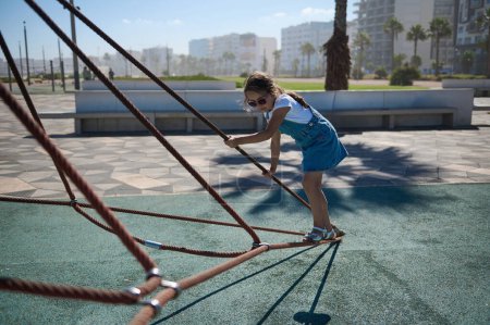 Photo for Little child girl spending active time climbing on the web in the city playground. Adorable little kid girl in casual denim and sunglasses, playing and having fun in urban amusement or adventure park - Royalty Free Image