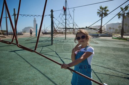 Photo for Smiling little girl in the city playground behind a climbing net. Adorable child in denim sundress, standing at the city playground behind a climbing net, looking at camera, through the sunglasses. - Royalty Free Image