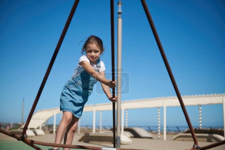 Little girl having fun in the adventure park. Adorable active child girl in casual denim sundress, climbing on the spider web and jumping on the trampoline during a weekend in the city promenade