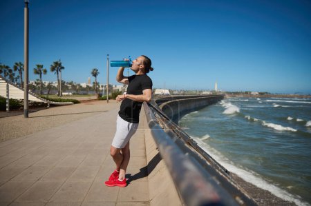Full length lifestyle portrait young athletic man, sportsman in activewear, drinking water after workout outdoor. Male athlete resting after sports training or morning jog on the Atlantic beach