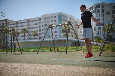Junger, athletischer Mann turnt mit einem Springseil auf dem städtischen Outdoor-Sportplatz. Ganzkörperporträt eines Sportlers, lateinamerikanischer Typ in aktiver Kleidung, der sich aufwärmt und durch ein Springseil springt