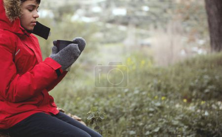 Close-up view of teenage boy in red parka and warm woolen mittens, holding a mug with hot drink, sitting on a log in the forest, relaxing while trekking and hiking in mountains on early spring day