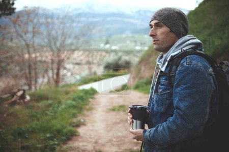 Téléchargez les photos : Homme de randonnée heureux aventurier détendu tenant une tasse thermos de voyage et regardant loin, debout sur le sentier forestier dans les montagnes. Beau jeune homme 40s rêveur regardant de côté tout en trekking dans la nature - en image libre de droit