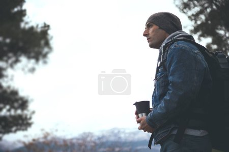 Téléchargez les photos : Portrait latéral d'un randonneur aventurier avec sac à dos, tenant une tasse thermos et regardant loin pendant la randonnée dans les montagnes de la Sierra de Cazorla. Personnes actives, trekking et concept de voyage - en image libre de droit