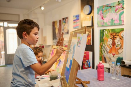 Foto de El talentoso niño de primaria se concentra en pintar cuadros sobre lienzos en aulas de arte creativo. Arte visual. Desarrollo de las habilidades artísticas de los niños. - Imagen libre de derechos