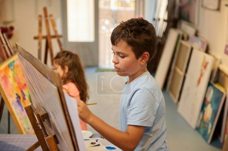 Foto de Retrato de confianza de un adolescente caucásico guapo en camisa casual azul, sosteniendo un pincel y dibujo sobre lienzo, mientras aprende arte en taller de arte creativo - Imagen libre de derechos