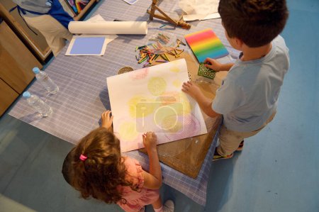 Foto de Vista desde arriba de un adolescente escolar y una colegiala de edad primaria dibujando sobre papel con lápices de colores pastel, creando un fondo para más durante la clase de arte en la escuela o galería de bellas artes. - Imagen libre de derechos