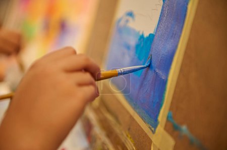 Foto de Primer plano de la mano de un estudiante sosteniendo pincel y pintura sobre lienzo durante la clase de arte visual en la escuela de arte - Imagen libre de derechos