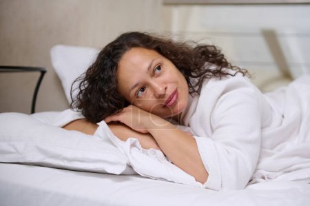 Close-up portrait of a beautiful curly haired woman in white bathrobe, dreamily looking away while relaxing, falling asleep on the white soft bed sheets on her comfortable bed in the bedroom