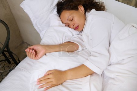 Overhead view of a multi ethnic young adult woman sleeping. High angle view of beautiful young woman in white pajamas, lying in bed and keeping her eyes closed while covered with white soft blanket.