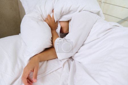 View from above of young woman covering her face with pillow while sleeping in a comfortable bed in the home bedroom in the early morning. It's time to wake up