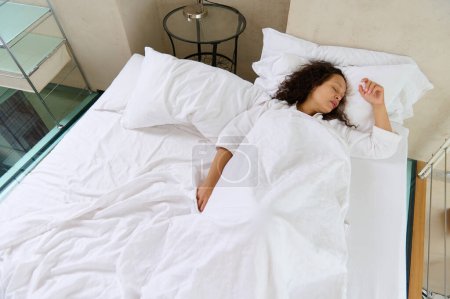 View from above of a multi ethnic woman in white pajamas, sleeping in the bed, covered with white soft blanket. Pretty female enjoying the softness and comfort of white pillow and bed sheets