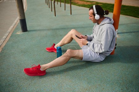 Young active athletic man in wireless headphones, checking mobile app on his smartphone while resting after workout the urban sports ground. Sportsman using mobile phone while working out outdoors