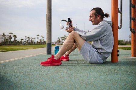 Young active athletic man checking mobile app on his smartphone while resting after workout the urban sports ground. Sportsman using mobile phone while working out outdoors. Side view