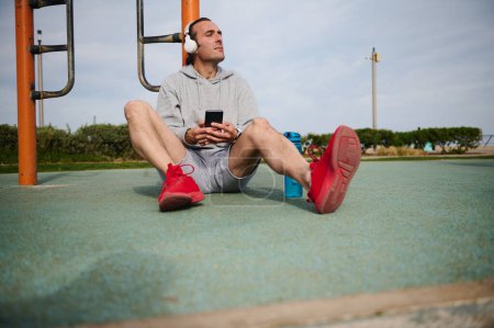 Young active athletic man listening music on wireless headphones, checking mobile app on his smartphone while resting after workout the urban sports ground. Active people, sport and modern technology