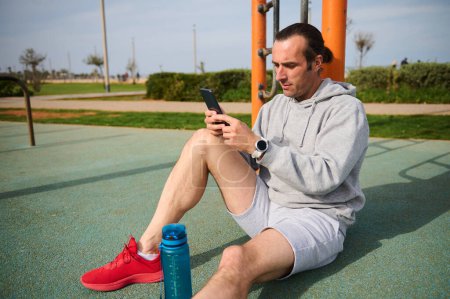 Young active athletic man checking health sport mobile app on his smartphone while resting after workout the urban sports ground. Active people, sport and modern technology