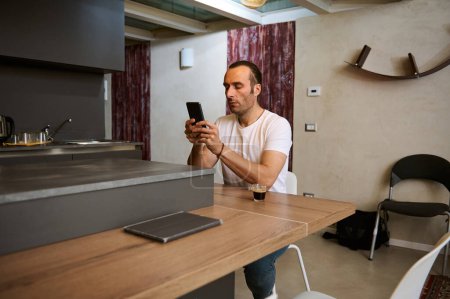 Attraktiver junger Mann checkt Handy-App auf seinem Smartphone, sitzt am Tisch in der heimischen Küche. Schöner Kaukasier mit Handy, Surfen im Internet und sozialen Medien und Spaß an modernen Technologien
