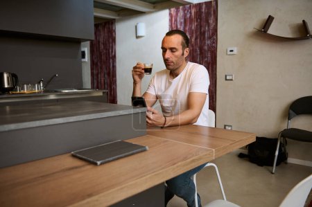 Porträt eines entspannten jungen Geschäftsmannes, der seinen morgendlichen Kaffee trinkt, Nachrichten auf seinem Smartphone liest, Social-Media-Inhalte checkt, im gemütlichen Wohnbereich am Tisch sitzt.