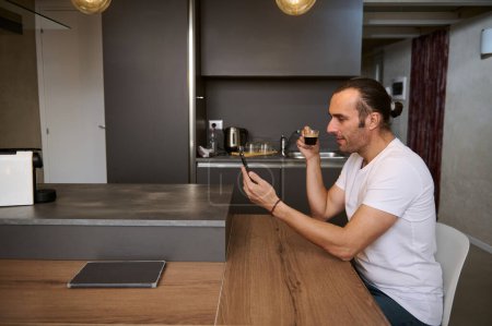 Seitenporträt eines entspannten jungen Geschäftsmannes, der seinen Morgenkaffee trinkt, Nachrichten auf seinem Smartphone liest, Social-Media-Inhalte checkt, am Tisch im gemütlichen Wohnbereich sitzt.