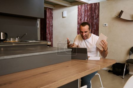 Stressed and worried man talking on mobile phone, arguing while unsatisfied by paying bills, doing paperwork, sitting at kitchen table at home interior. Young guy expressing anger planning budget