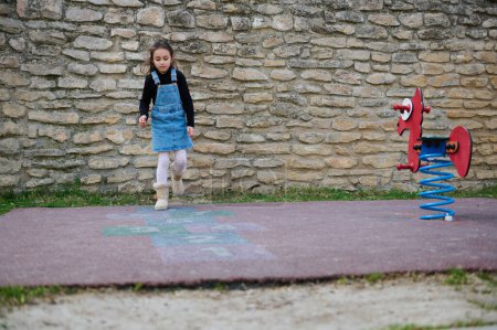 Photo for Full length shot of a lovely little child girl playing hopscotch on the school playground. Beautiful kid in denim sundress enjoying active leisure games outdoors, jumping on squares, playing hopscotch - Royalty Free Image