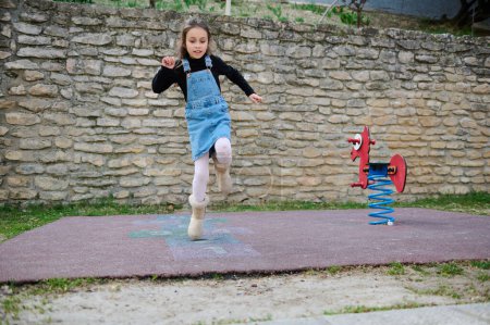 Photo for Full length portrait of a little kid girl jumping on one leg, playing hopscotch on a playground outdoors. Hopscotch popular street game. Active children. Happy carefree childhood. People and lifestyle - Royalty Free Image