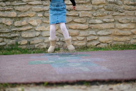Photo for Cropped view. Focus on legs of a child girl playing hopscotch on the school playground. Beautiful kid in denim sundress enjoying active leisure games outdoors, jumping on squares, playing hopscotch - Royalty Free Image
