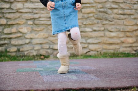 Photo for Details on the legs of an active little child girl playing hopscotch, takes turns jumping over squares marked on the ground. Street children's games in classics. Childhood. Kids and healthy lifestyle - Royalty Free Image