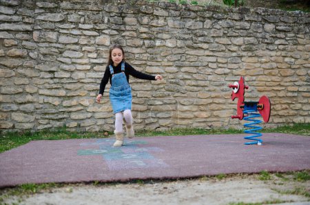 Photo for A sporty 6-7 years old kid girl playing hopscotch, taking turns jumping over the squares marked on the school play ground. Street children's games in classics. Childhood and healthy lifestyle concept - Royalty Free Image