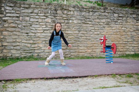 Photo for Active little child girl, elementary age schoolgirl plays hopscotch, takes turns jumping over squares marked on the ground. Street children's games in classics. Childhood. Kids and healthy lifestyle - Royalty Free Image