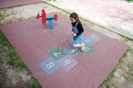 Photo for View from above of a kid girl playing hopscotch, taking turns jumping over the squares marked on the school play ground. Street children's games in classics. Childhood and healthy lifestyle concept - Royalty Free Image