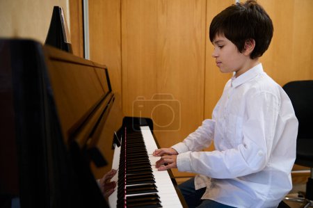 Confident teenage pianist plays piano at home, sitting at pianoforte, dressed in white shirt. Authentic portrait of handsome boy musician performing classic melody, practicing chord musical instrument